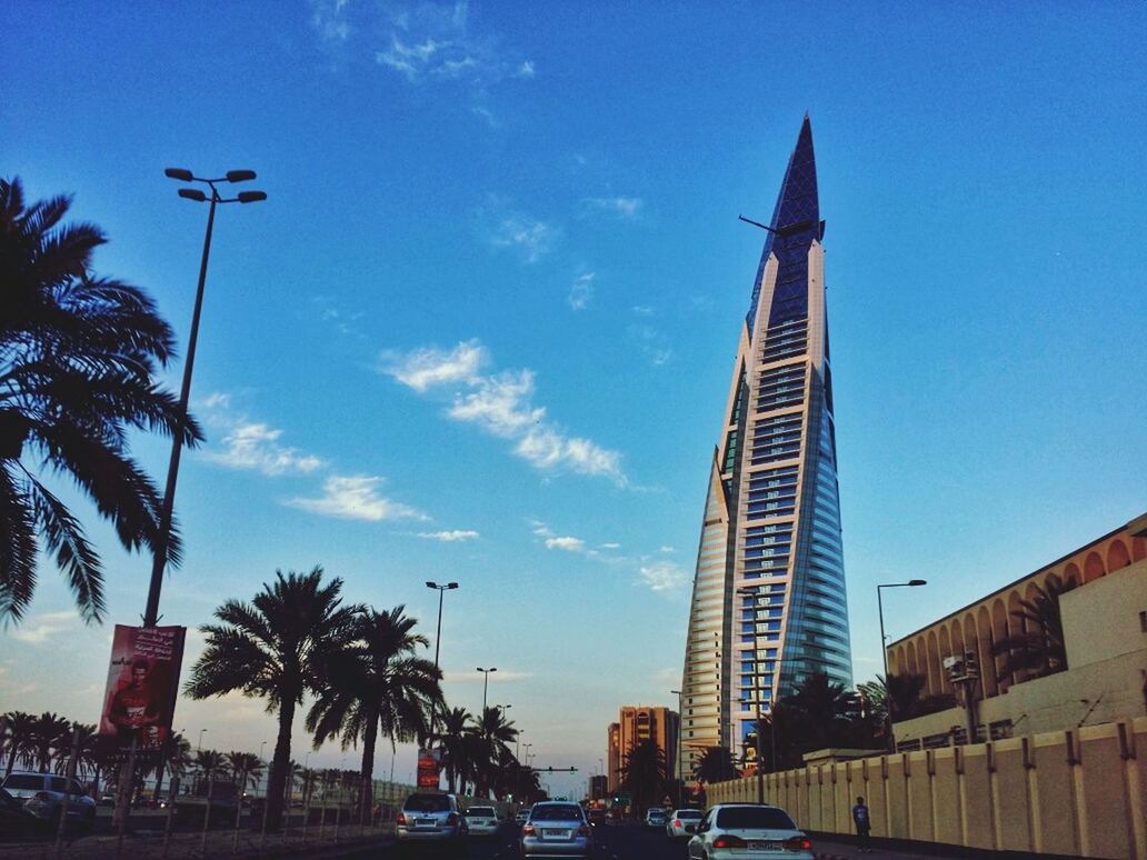 building exterior, architecture, built structure, car, city, land vehicle, transportation, tall - high, low angle view, tower, blue, sky, mode of transport, tree, street, road, skyscraper, modern, incidental people, street light