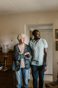 Happy male caretaker walking arm in arm with senior woman in living room at home