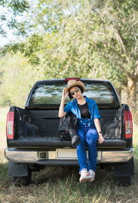 Full length of woman wearing hat sitting on car trunk outdoors