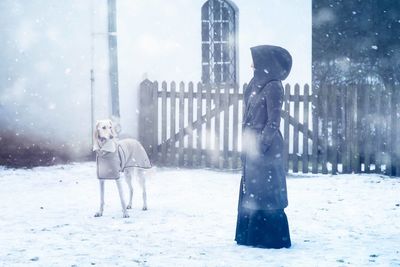 Woman wearing hooded coat with dog against house during snowfall