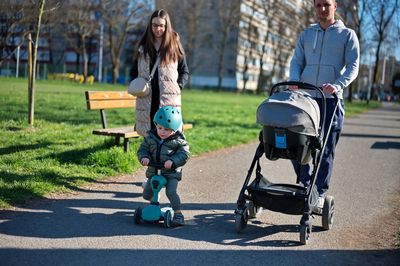 Little toddler on scooter and his parents with baby stroller walking in a park