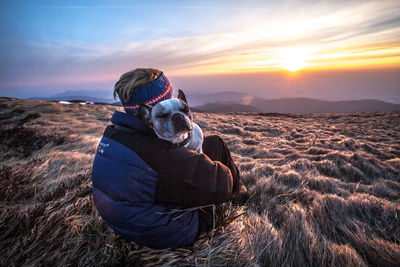 Man with dog sitting on grass against sky during sunset