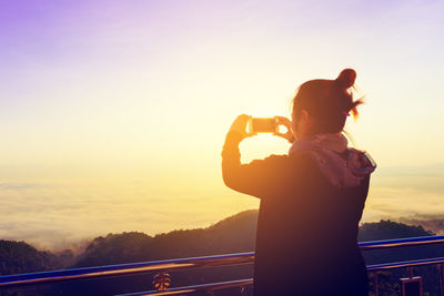 Rear view of woman photographing against sky during sunset