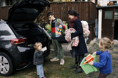 Parents with children loading luggage in electric car trunk at back yard