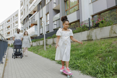 Girl skateboarding with mother and father walking in background at footpath