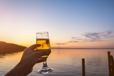 Hand holding glass of water against sky during sunset