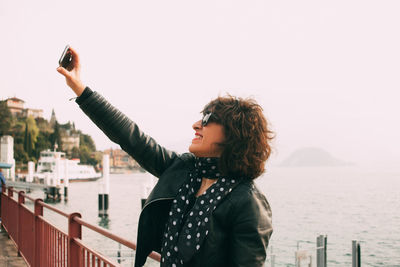 Side view of smiling woman taking selfie by sea against clear sky