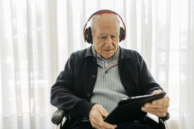 Senior man sitting in wheelchair reading using digital tablet while listening music with headphones