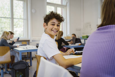 Side view portrait of smiling schoolboy sitting at desk in classroom