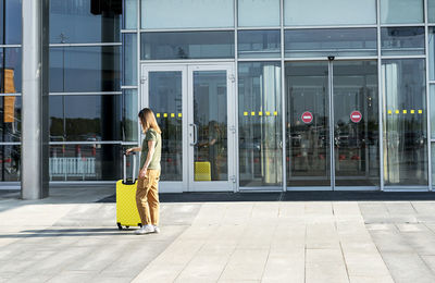 Young woman traveler carrying a yellow suitcase next to the entrance to the airport outside luggage