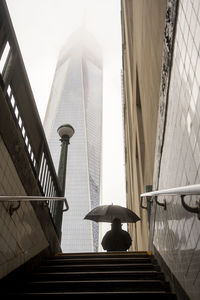 Usa, new york, new york city, pedestrian standing with umbrella in front of subway entrance with skyscraper shrouded in fog in background
