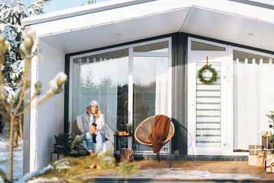 Smiling woman with blonde in winter clothes with coffe cup sitting near country house in winter