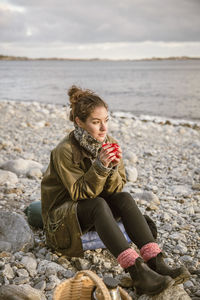 Thoughtful woman sitting on rock with drink at beach