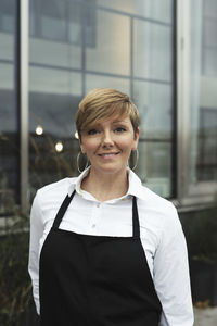 Portrait of smiling professional wearing apron standing against glass building