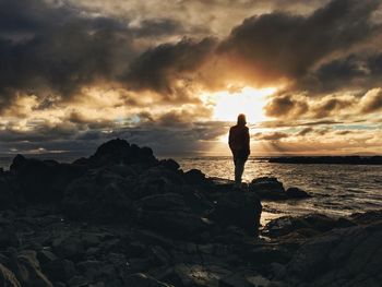 Silhouette woman standing on rock by sea against sky during sunset