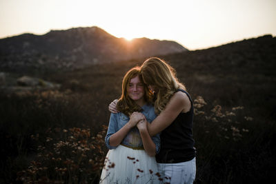 Mother embracing daughter while standing on field against mountains during sunset