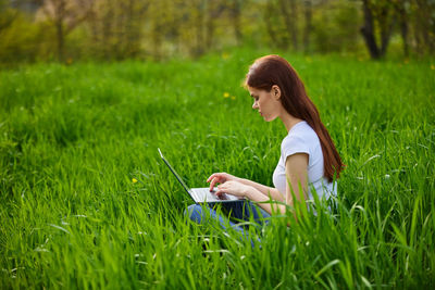 Side view of young woman using laptop while sitting on grassy field