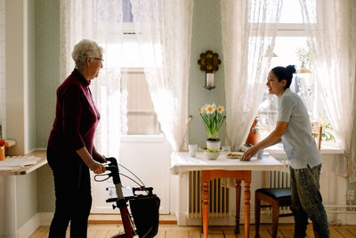 Smiling young female caregiver serving breakfast while looking at retired senior woman walking with rollator in nursing