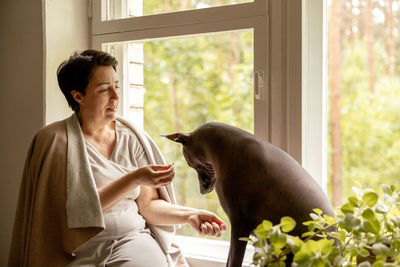 Middle age beautiful woman sitting on windowsill with her dog. 50-year-old woman