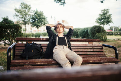 Thoughtful man with hands behind head sitting on wooden bench against sky at park