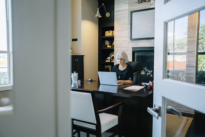 Businesswoman working on laptop computer at desk in office seen through doorway