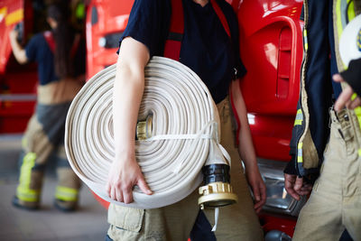 Midsection of female firefighter holding rolled up fire hose while standing with coworker at fire station