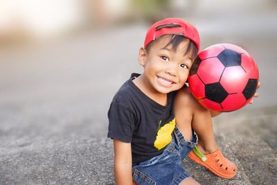Portrait of cheerful boy sitting with ball on rock