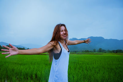 Young woman with arms outstretched standing on field against sky