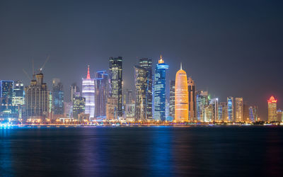 Illuminated buildings by sea against sky at night