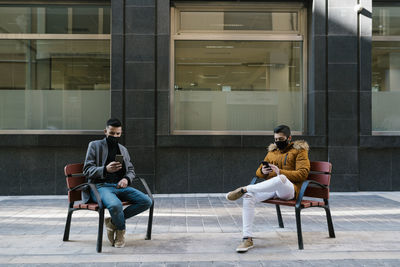 Young men wearing face mask using mobile phones while sitting on chair in city