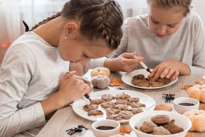 Two girls decorate halloween gingerbread cookies on plates with chocolate icing. 