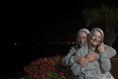 Portrait of man embracing woman while sitting against sky at night