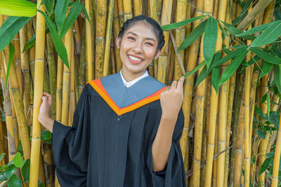 Portrait of smiling young woman in graduation gown standing against bamboo