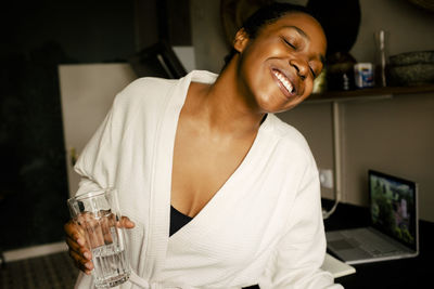 Cheerful young woman holding drinking glass while dancing in kitchen at home