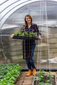 A young woman is smiling standing in a greenhouse holding a tray with seedlings of vegetables