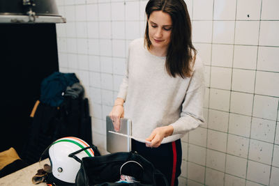 Young busineswoman removing laptop from bag while standing against wall at small office