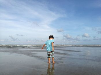 Rear view of boy standing on shore at beach against cloudy sky