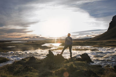 Rear view of woman standing on rocks by river against sky on sunny day