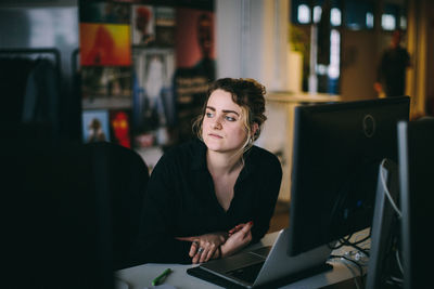 Young woman looking away while sitting by laptop on table at office