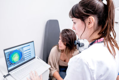 From above of focused female ophthalmologist using laptop with eyesight check up results during appointment with patient in modern hospital