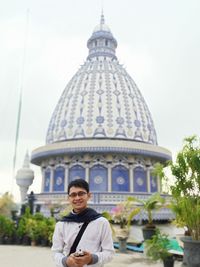 Portrait of young man holding mobile phone against building and sky