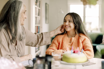 Senior woman touching cheek of granddaughter sitting with birthday cake at dining table