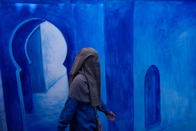 Woman wearing a niqab walks in front of a blue painted mural in ancient medina. chefchaouen. morocco