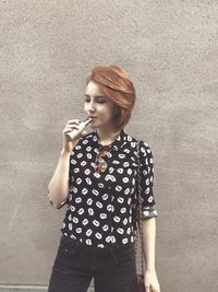 Beautiful young woman smoking while standing against wall