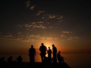 Silhouette people fishing in sea against orange sky during sunset