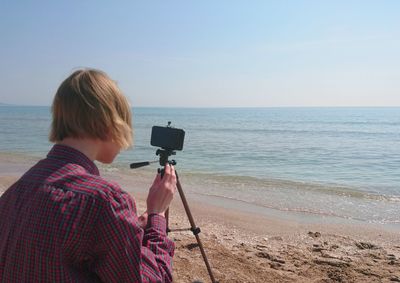 Rear view of woman photographing on beach against sky