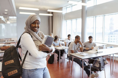 Portrait of smiling student in hijab standing with book against friends in classroom