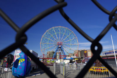 Ferris wheel by chainlink fence against sky in city