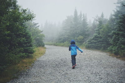 Rear view of boy wearing hood and running on road amidst trees in foggy weather