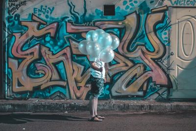 Side view of woman holding balloons over face while standing against graffiti wall